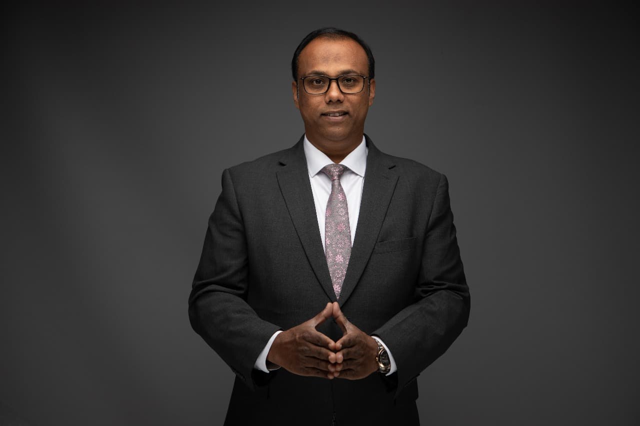 Renoy standing in a dark suit against a charcoal studio background